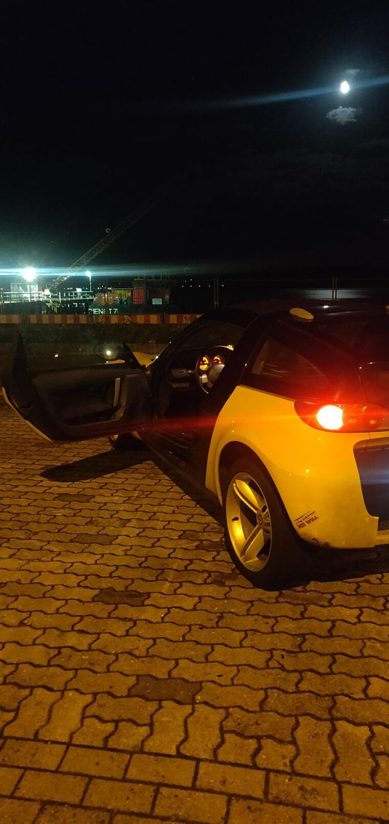 Gelber Smart Roadster bei Nacht von hinten, Mond über dem Wasser